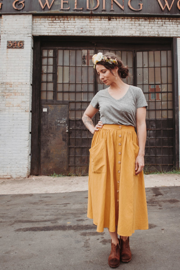 Meg wears a yellow estuary skirt and a grey t-shirt and a flower crown. She stands in front of an old door on an old building
