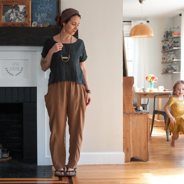 Meg wearing a charcoal strata top, ochre arenite pants, standing next to a fireplace