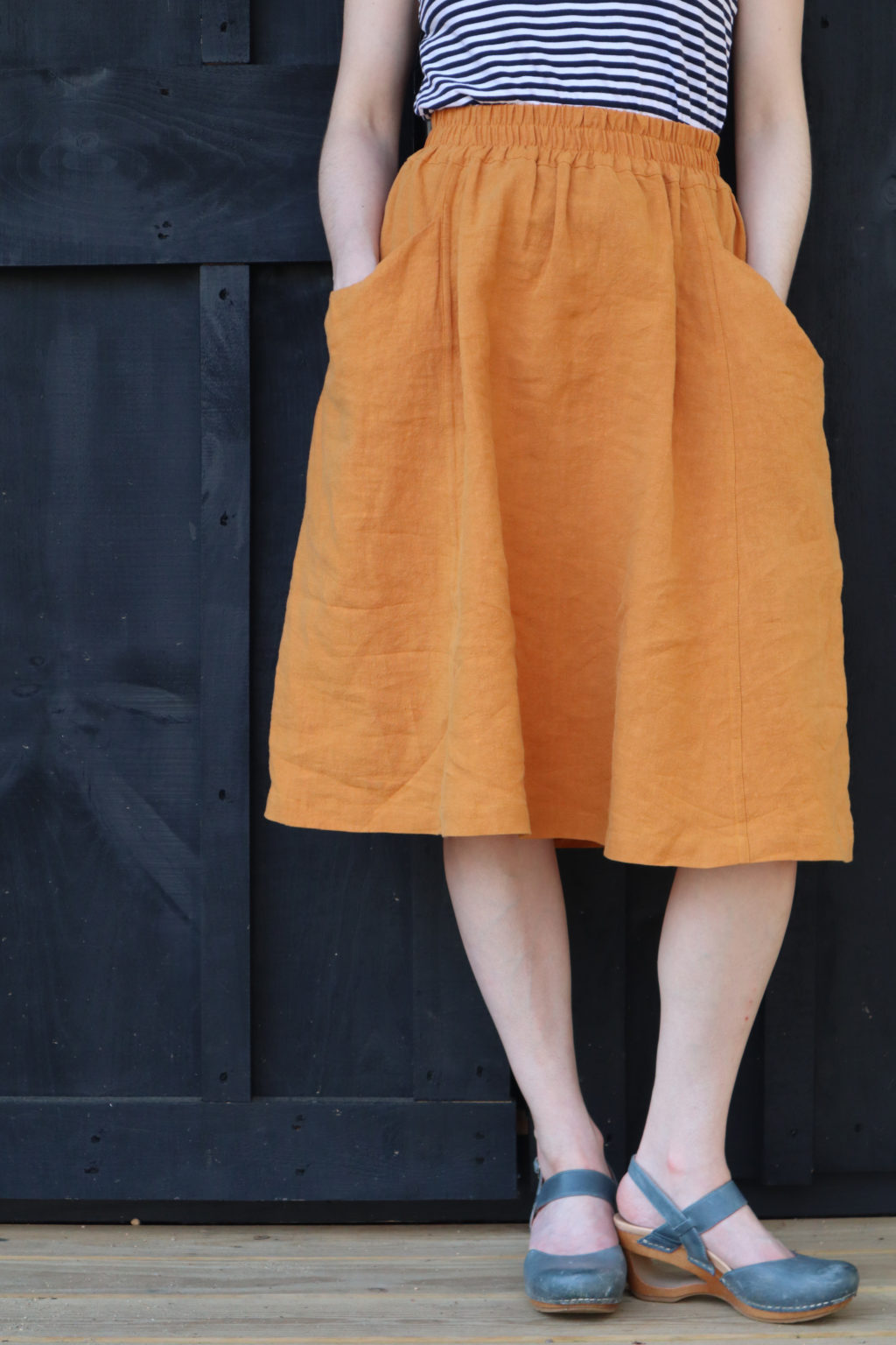 a waist down shot of an orange view A gypsum skirt worn with blue clogs and s triped shirt, against a black wall