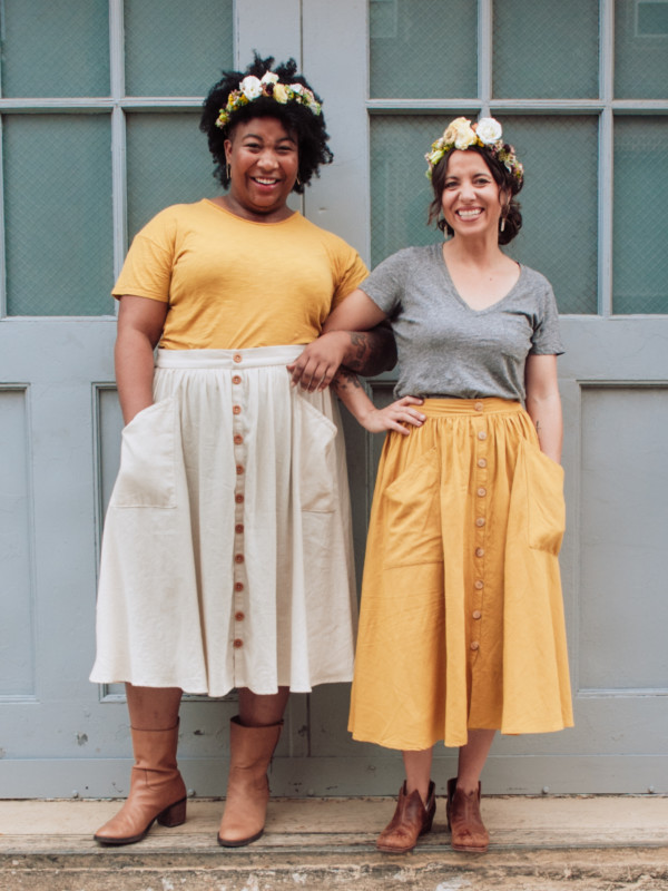 Meg and Ashley stand against a grey door wearing Estuary skirts, t-shirts and flower crowns
