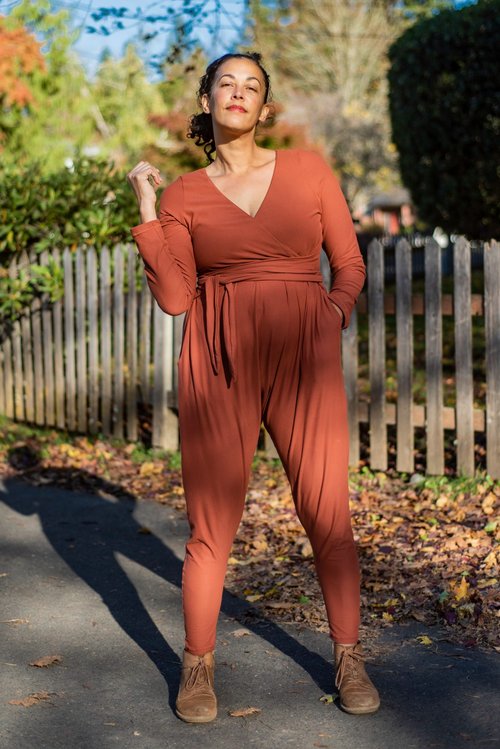 Ambrosia wears a brick red jumpsuit standing near a wooden fence. 