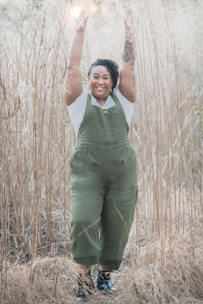 Ashley wears green overalls and a white short sleeved shirt. She's standing among tall grasses, withe her arms in the air, smiling at the camera. 