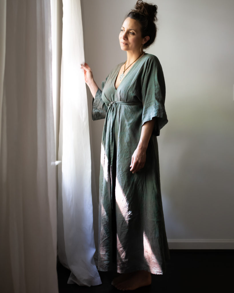 Meg wears a soft blue-gray floor length dress, standing by a window with white curtains. 
