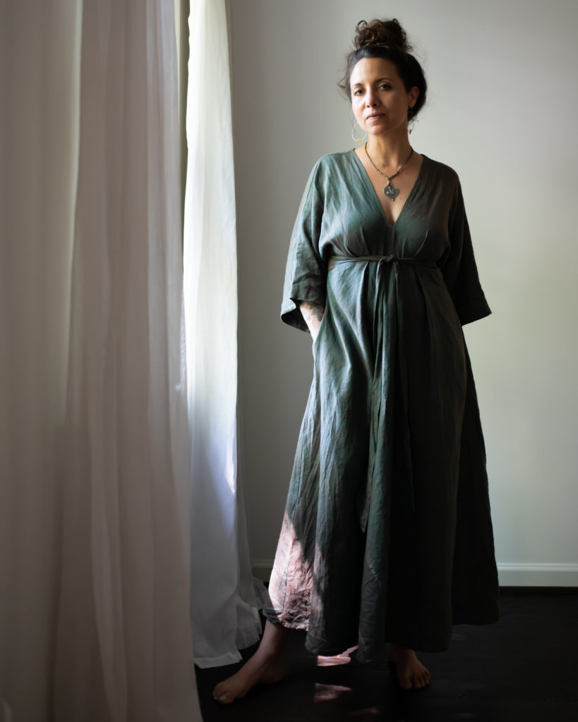 Meg wears a soft blue-gray floor length dress, standing by a window with white curtains. 