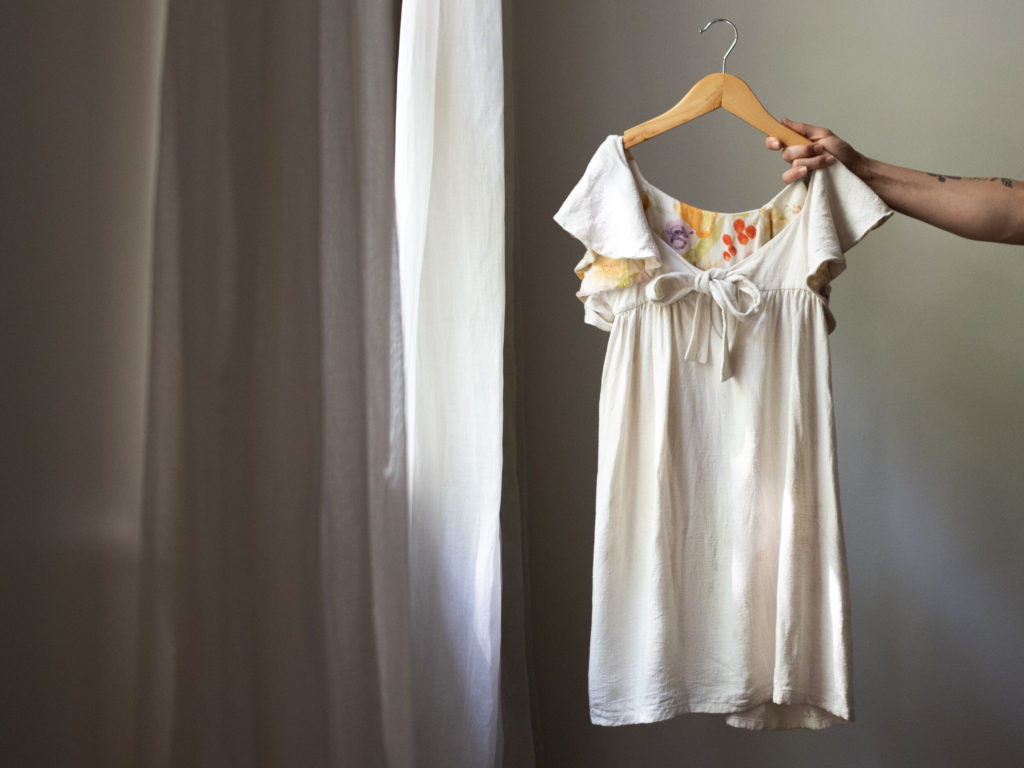 Meg holds a cream flower girl dress with a floral lining on a wooden hanger by a window with white curtains. 