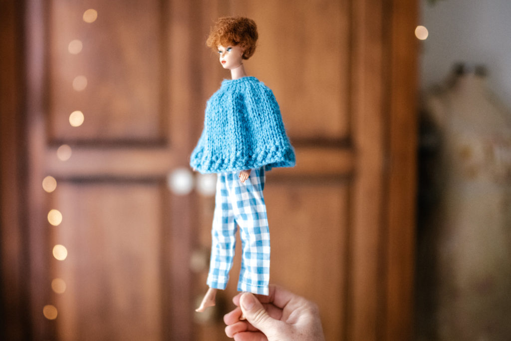 Megs hands holding a Vintage barbie wearing a blue sweater and matching blue gingham pants with a wooden wardrobe and fairy lights blurred in the background. 