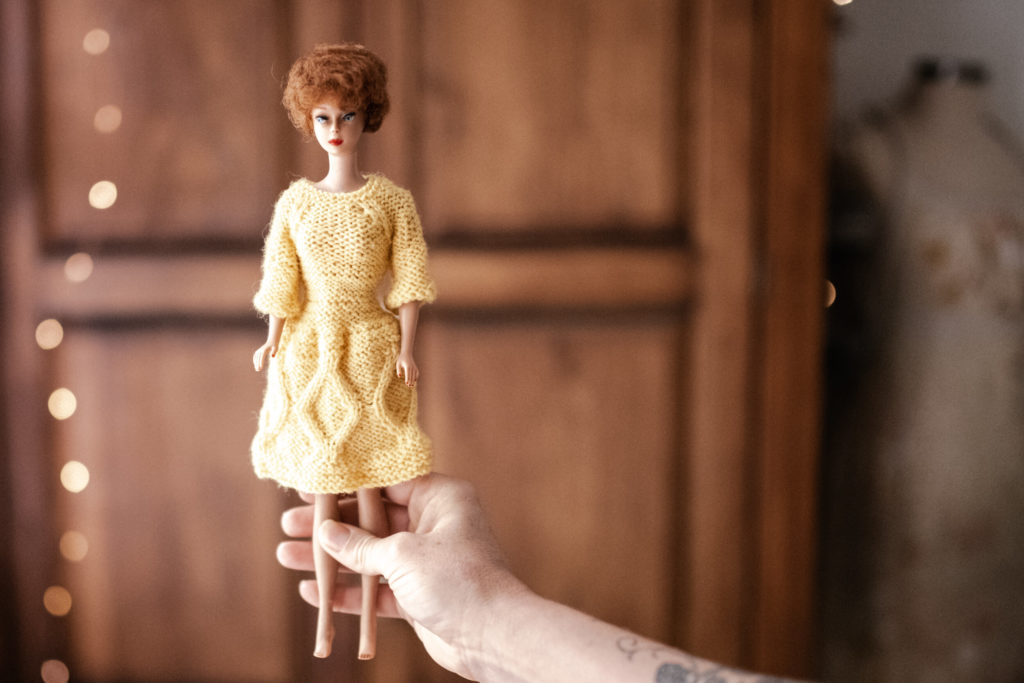 Megs hands holding a Vintage barbie wearing a denim skirt and red striped top with a wooden wardrobe and fairy lights blurred in the background. 