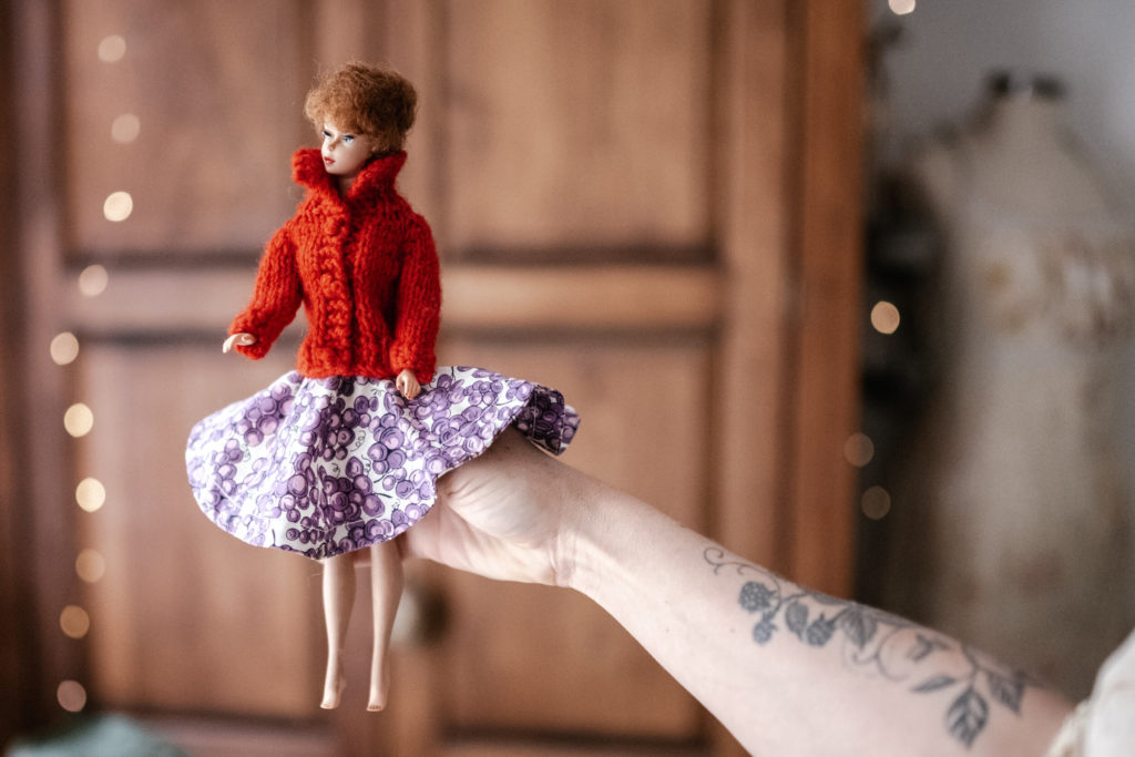 Megs hands holding a Vintage barbie wearing a red sweater coat and purple and white floral skirt with a wooden wardrobe and fairy lights blurred in the background. 