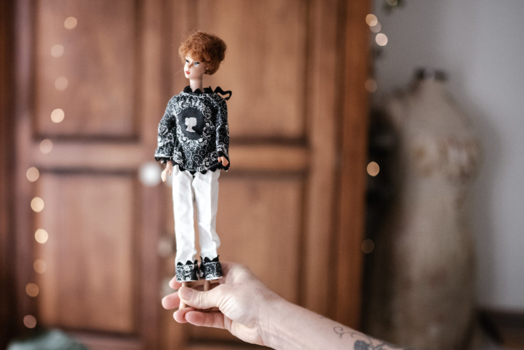 Megs hands holding a Vintage barbie wearing white pants trimmed in a black 70s print and a bloack boho style shirt with a wooden wardrobe and fairy lights blurred in the background. 
