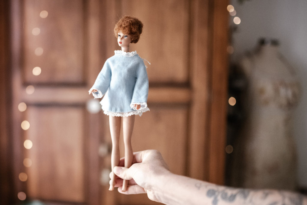 Megs hands holding a Vintage barbie wearing a satin blue minit dress with lace hem with a wooden wardrobe and fairy lights blurred in the background. 
