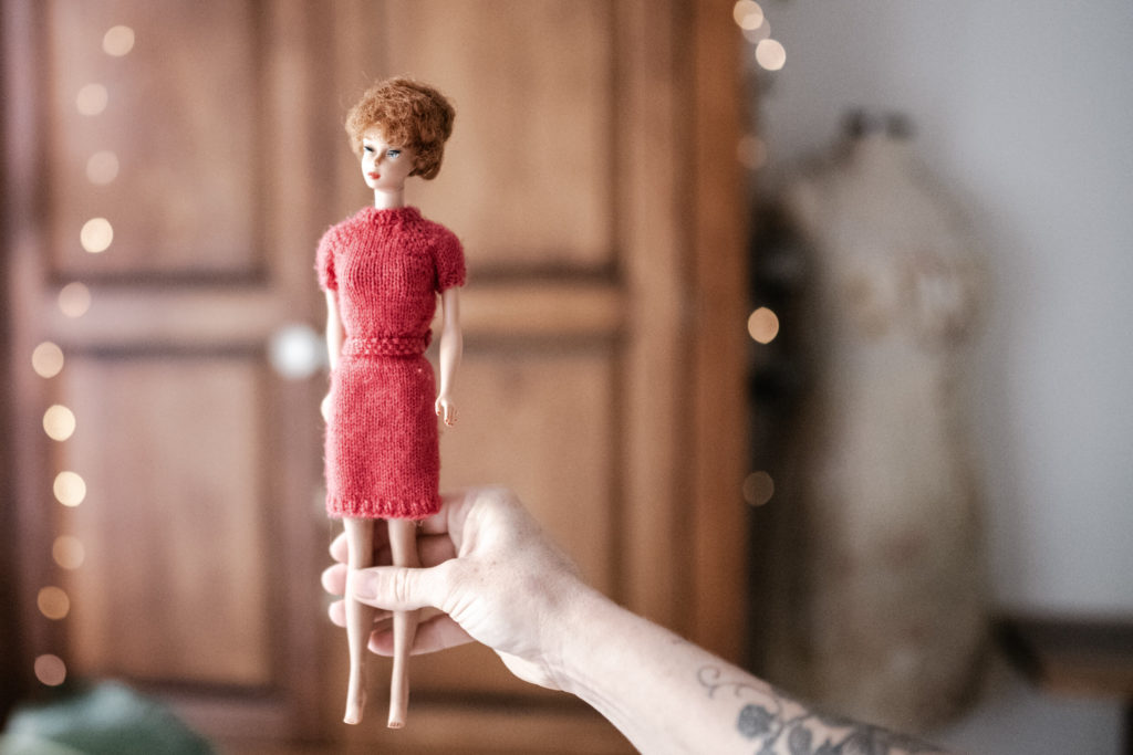 Megs hands holding a Vintage barbie wearing a red sweater and skirt set with a wooden wardrobe and fairy lights blurred in the background. 