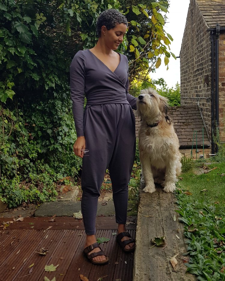 Emily wears a grey jumpsuit and stands next to her dog, who is perched on a wall. 