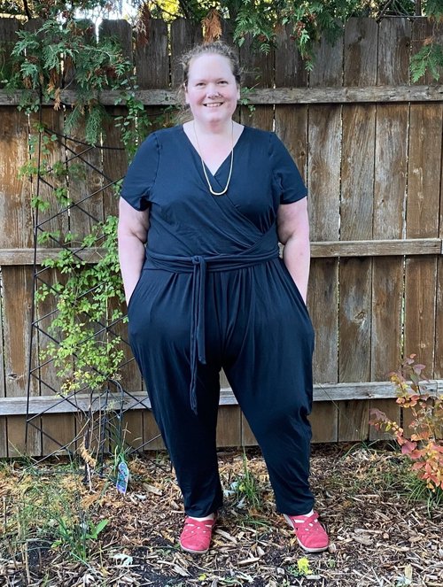 Emily wears a black jumpsuit standing in front of a wooden fence. 