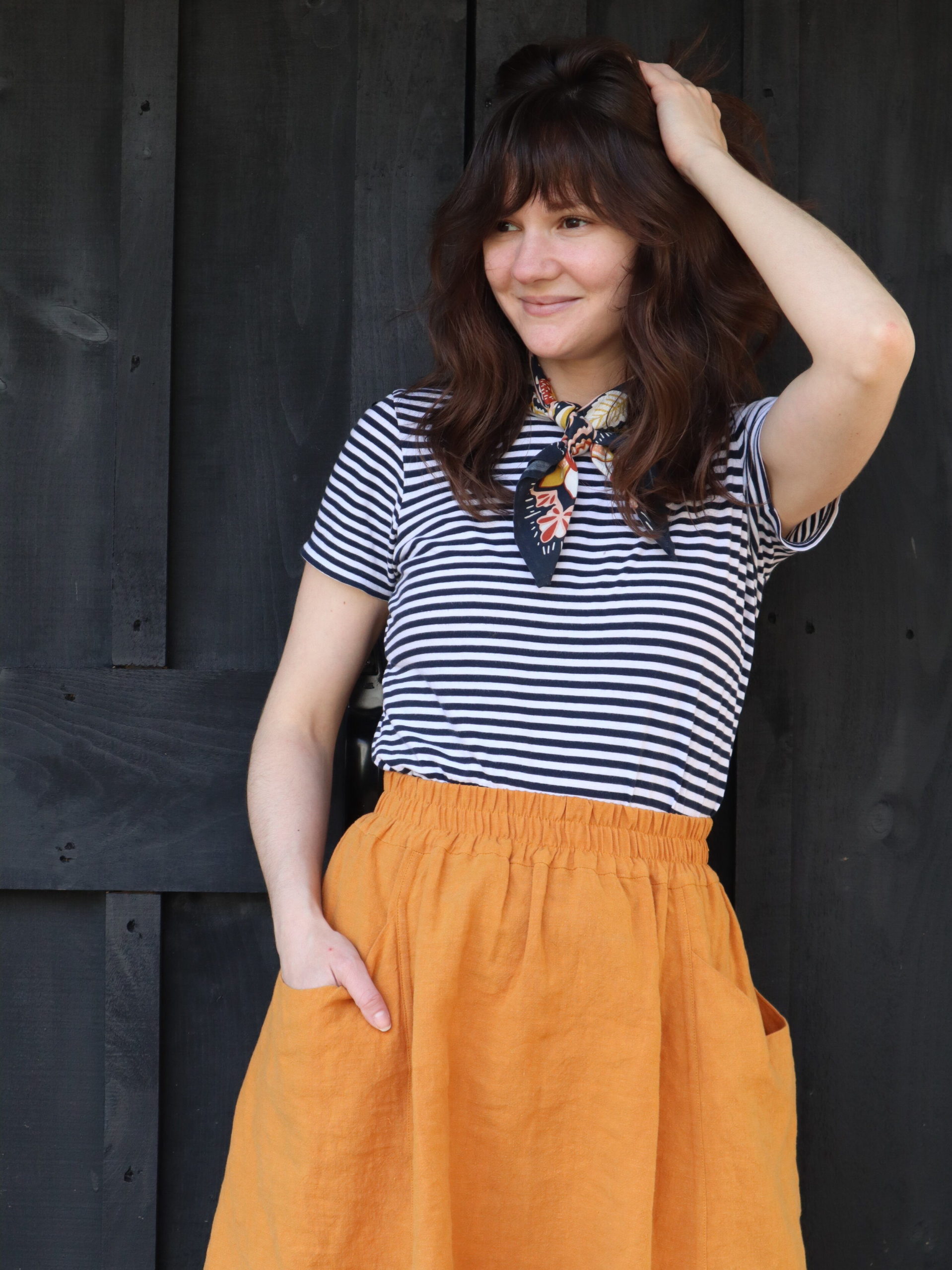 Meredith wears a striped tee and orange gypsum skirt in front of a black wooden wall