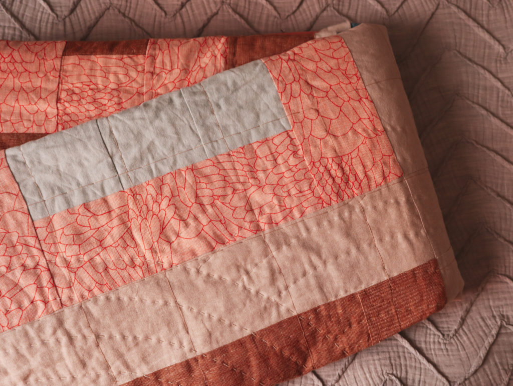 a beige, coral, blue and brown pieced quilt with hand quilting in progress on a grey duvet cover