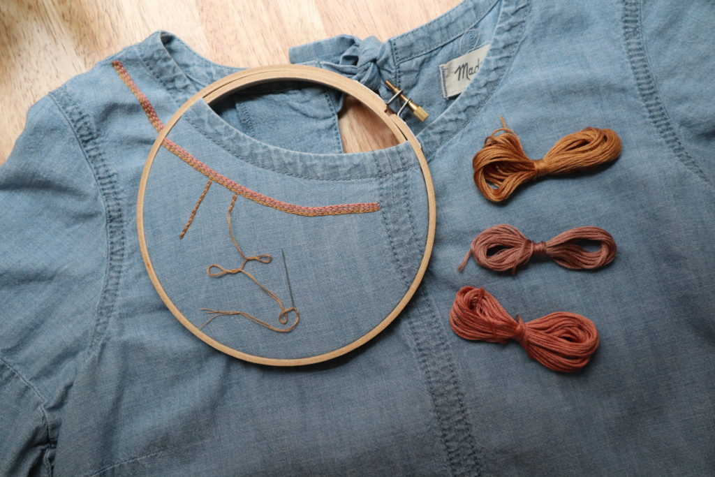 a denim shirt with an embroidery hoop place near the collar and chain stitching in progress in pink, gold, and mauve. 