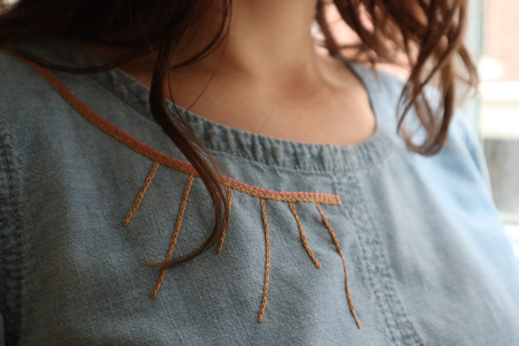 a close up of Meredith wearing a denim shirt with chain stitch embroidery of a sun-like geometric design, with one lock of hair falling over the embroidery. 