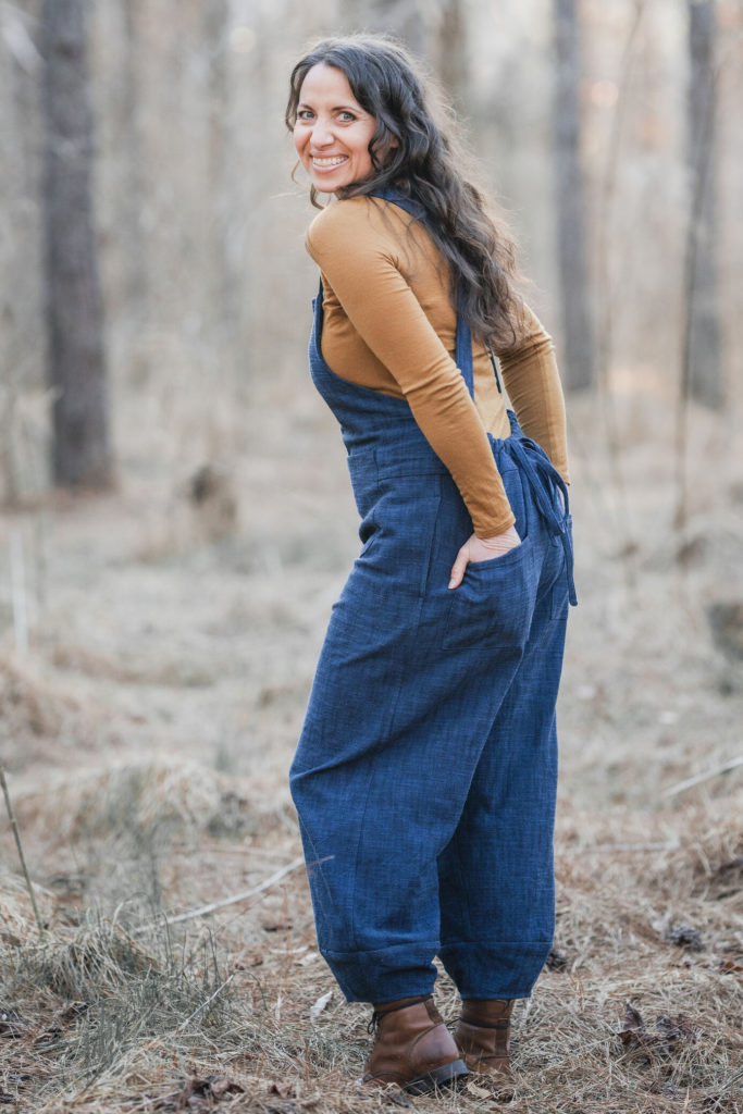 Meg stands in the woods looking over her shoulder to smile at the camera with her hands in her back pockets. She wear denim overalls and an ochre shirt. 
