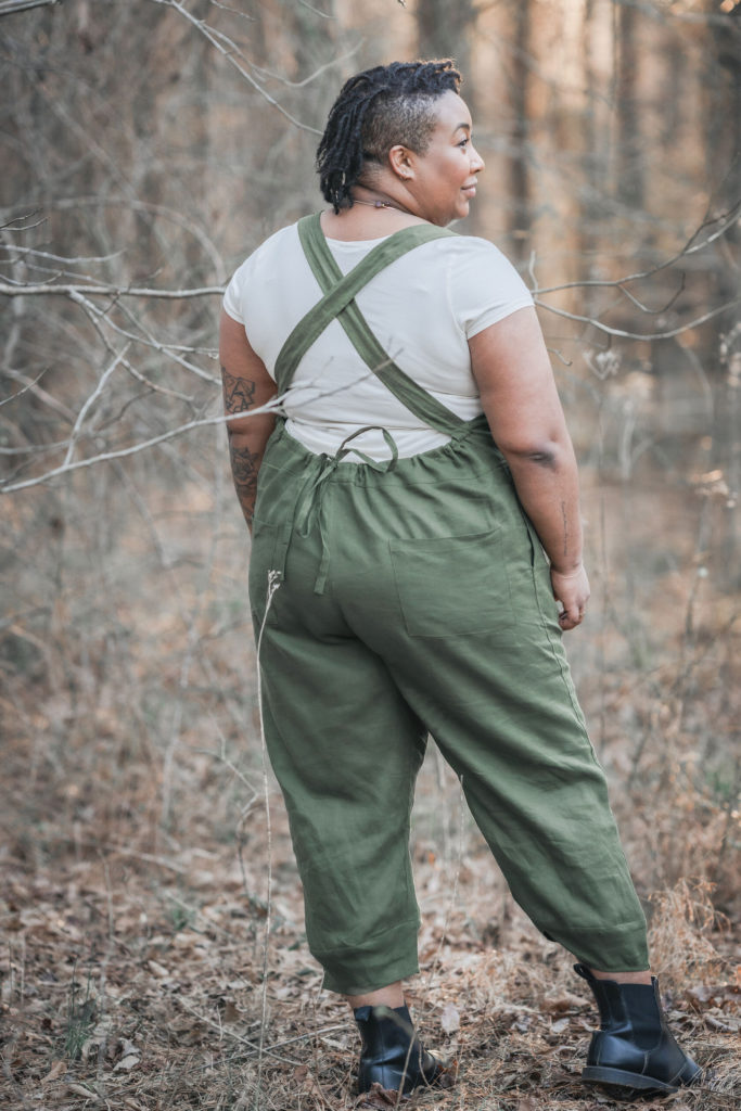 Ashely faces away from the camera, standing in the woods wearing green overalls and a cream shirt. She smiles and looks to the side 