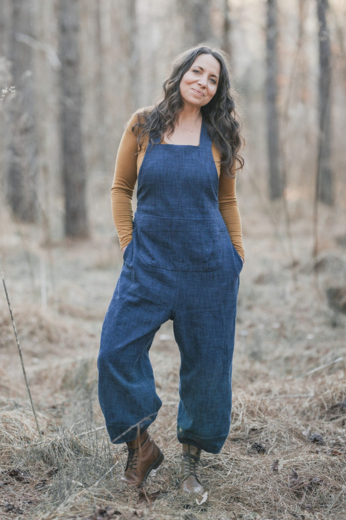 Meg wears denim overalls and an ochre long sleeved top. She standing in the woods, smiling at the camera with her hands in her pockets. 