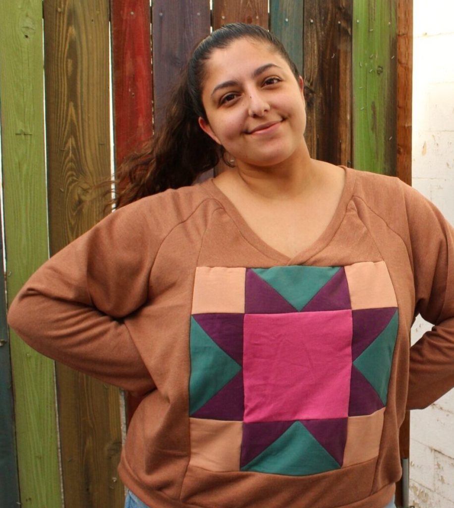 Romy wears a brown, tan, purple, teal, and pink nest sweatshirt with the quilt block motif