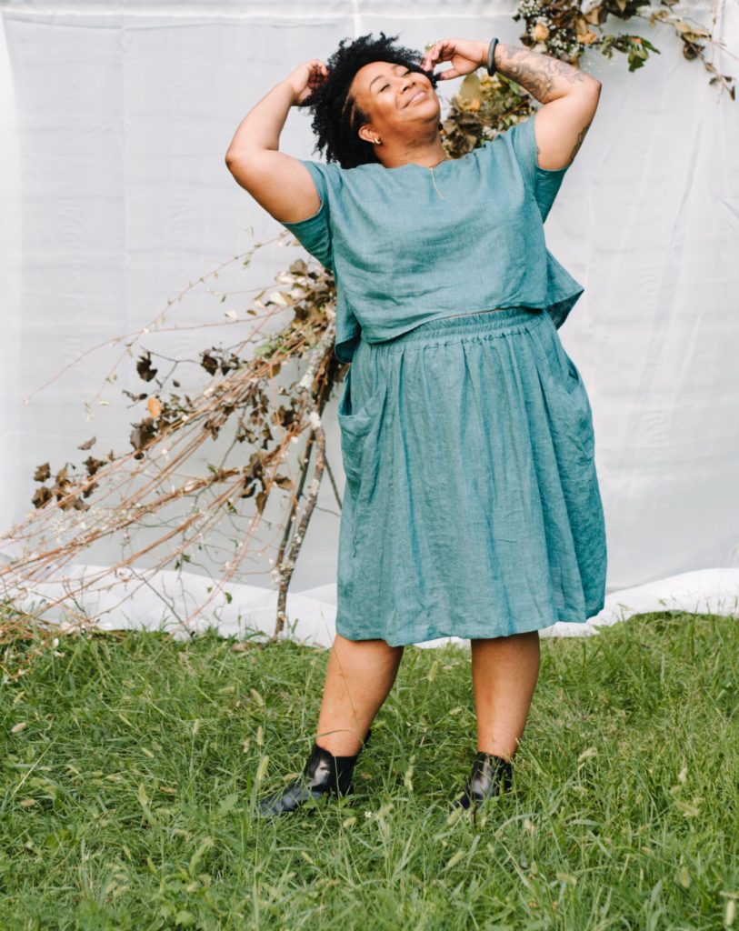 Ashley wears a matching teal blue strata top and gypsum skirt and stands in front of a white backdrop draped in flowers. She is smiling with her eyes closed and face raised towards the sky and her hands in her hair. 