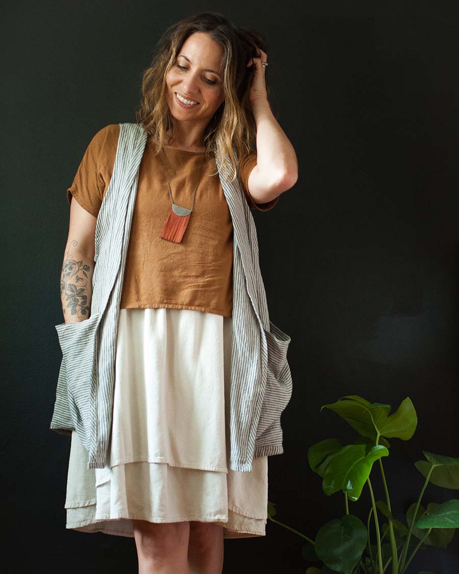 Meg wears a Forager Vest over an ochre colored Strata Top and white dress