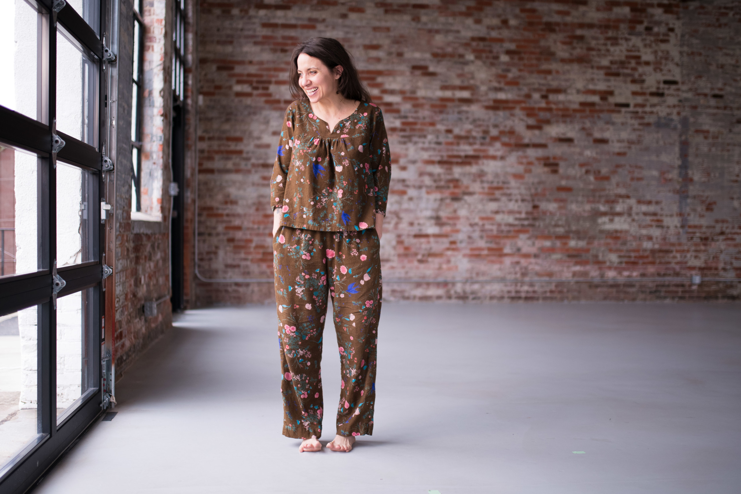 Meg is wearing brown floral, flannel Nocturne Pajamas in front of a brick wall.