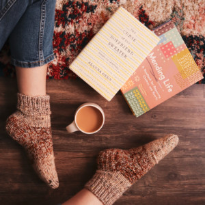 A person sits on the floor, with only her handmade socks, a cup of tea, and two books on visible.