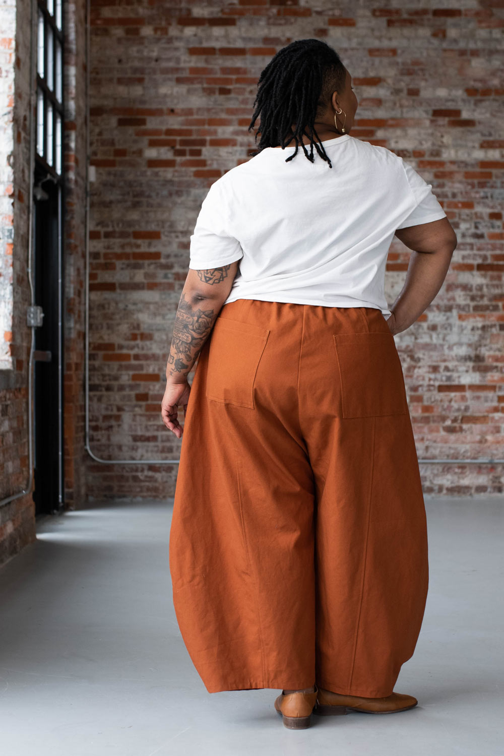 Ashley is wearing pumpkin orange Arthur pants and a white tee. She is facing away from the camera in front of a brick wall.