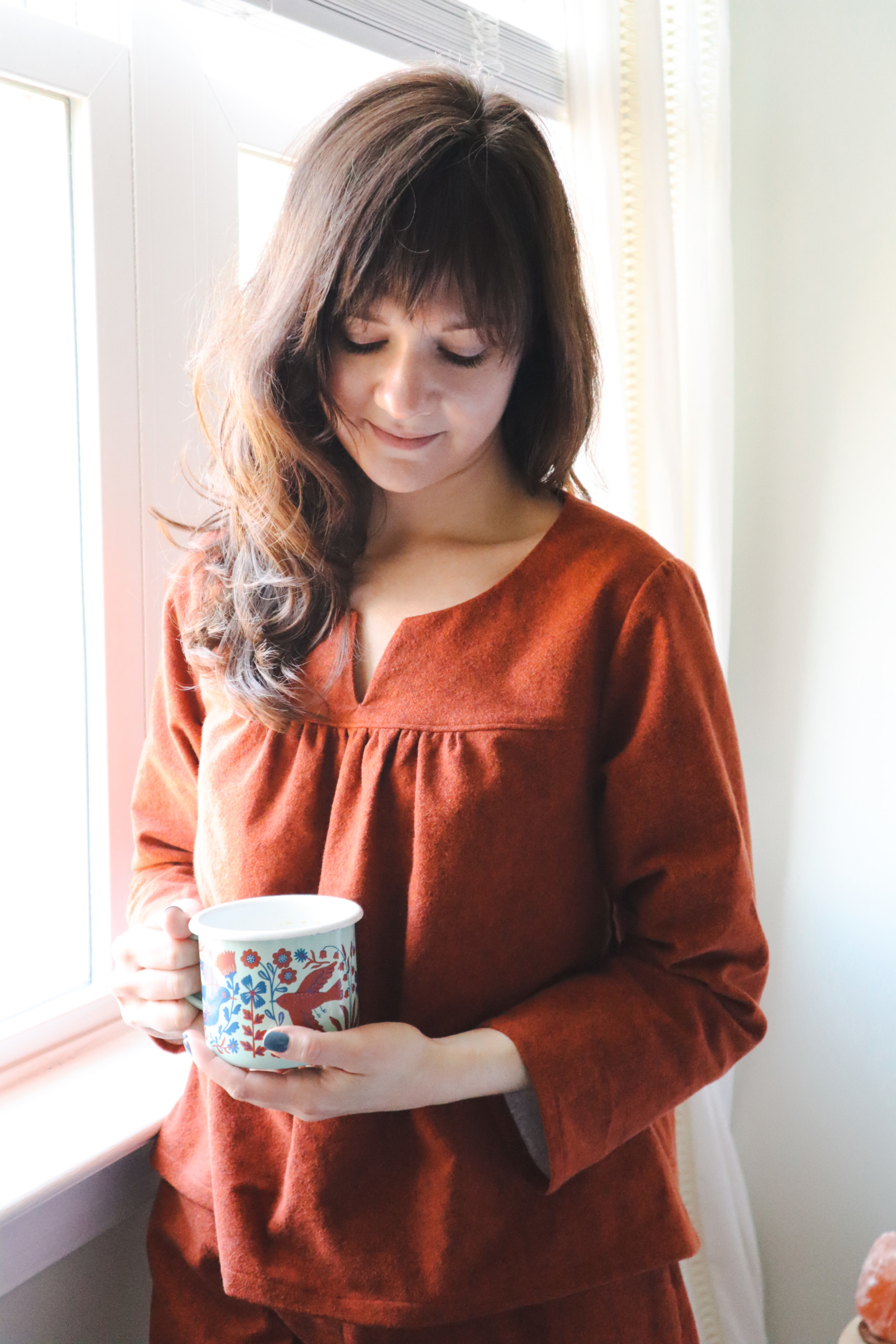 Meredith stands by a window wearing a red flannel nocturne top and holding a mug of coffee
