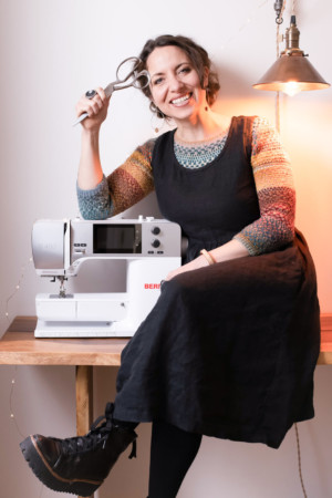 Meg wears a black, sleeveless dress over a colorful handknit sweater. She is sitting next to her sewing machine and holding scissors in her hand.