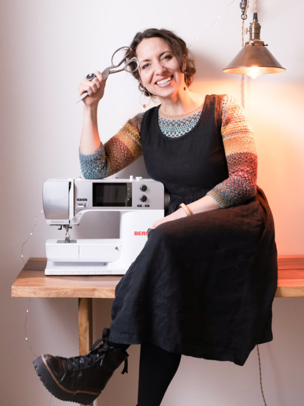 Meg wears a black, sleeveless dress over a colorful handknit sweater. She is sitting next to her sewing machine and holding scissors in her hand.
