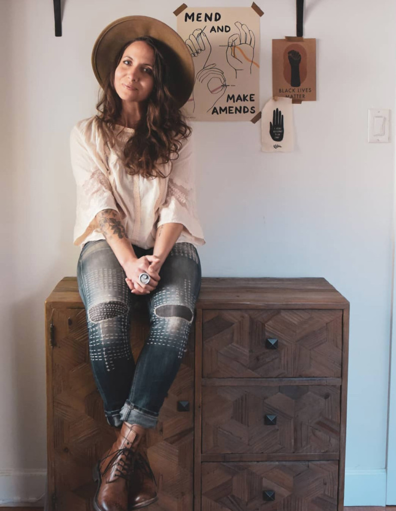 Meg sits on a wood cabinet. She is wearing mended jeans and a thrifted top. There is a poster on the wall behind her that says "Mend and make amends."