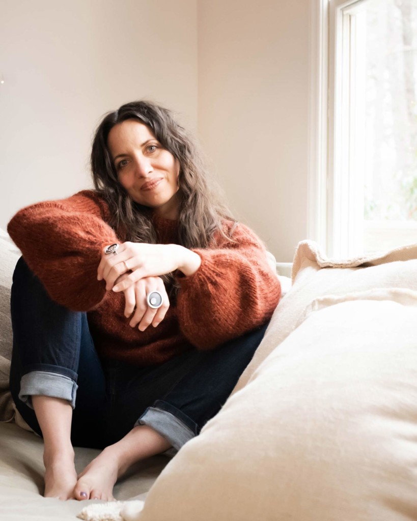 Meg sits on a sofa next to a window with her legs folded up beneath her arms. She is wearing a soft handknit rust sweater with voluminous sleeves and cuffed handmade jeans.