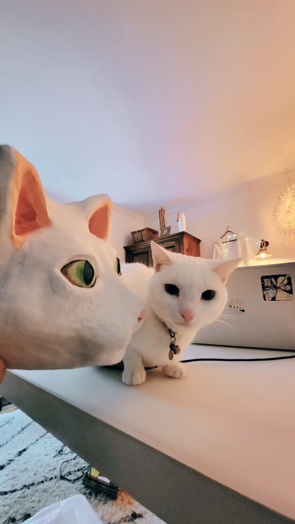 A real white cat sits, unnerved, next to a large paper mache mask replica of herself.