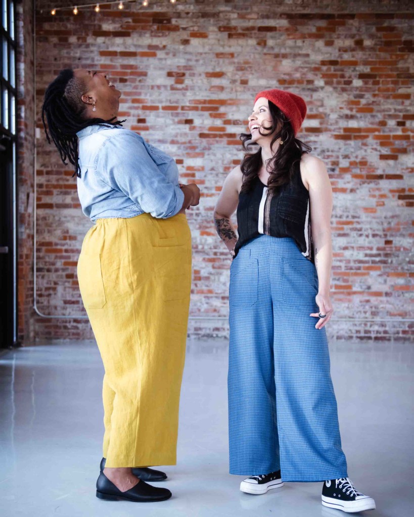 Showing fabric for cosecha, Ashley and Meg laugh together. Ashley wears yellow Cosecha pants and a chambray shirt, Meg wears blue checked Cosecha pants and a black tank and a red hat. 