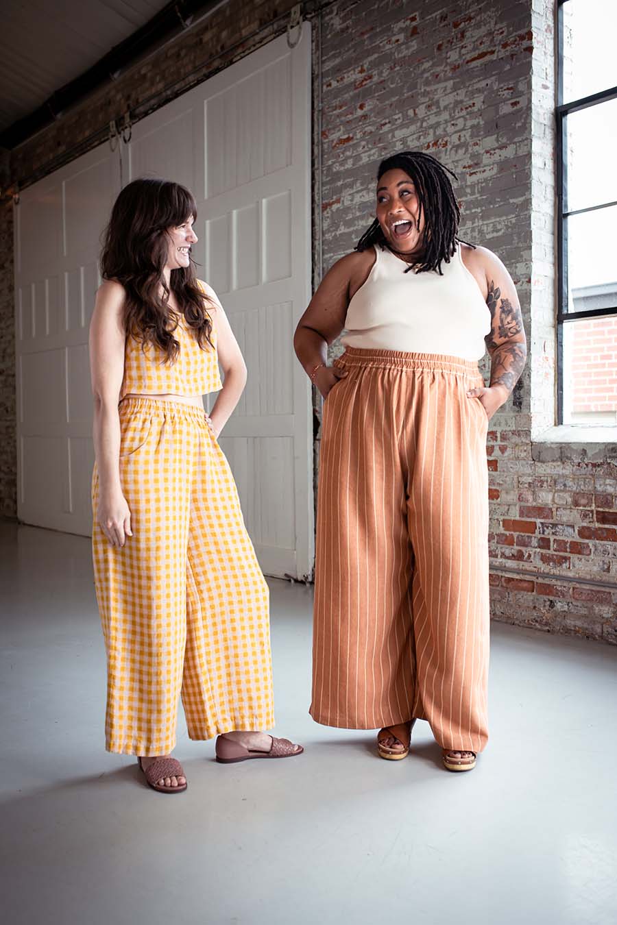 Ashley wearing striped view a chanterelle pants and a cream tank. Meredith wears pink and yellow checked matching separates. They smile and laugh together.