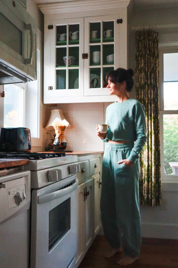 Meredith wears teal Chanterelle joggers and a matching sweatshirt, holding a cup of coffee in the kitchen. 